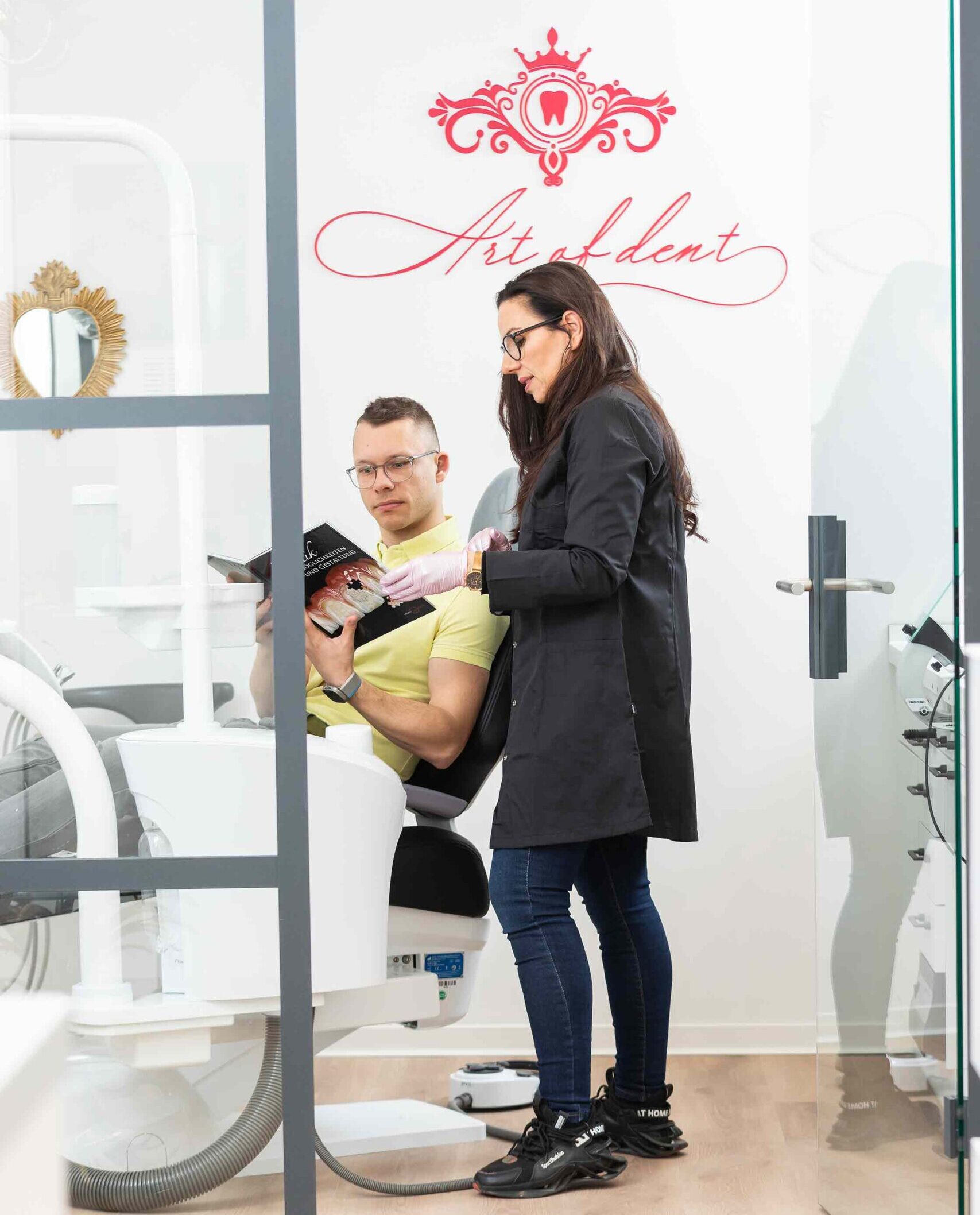 Beratung-Art-of-dent-Dentallabor-Zahntechnik-Potsdam-Babelsberg-Jenny-Ulrich Zahntechnikerin von Art of dent in Potsdam Babelsberg berät einen Patienten mit einem Buch zu Zahnersatz-Beispielen im Behandlungsraum.